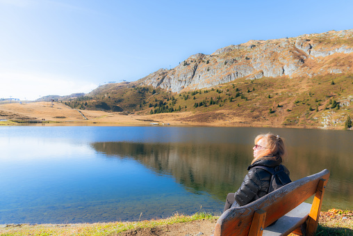Relaxed woman sitting on a wooden bench while enjoying the nature scenery on a sunny autumn day. Travel concept.