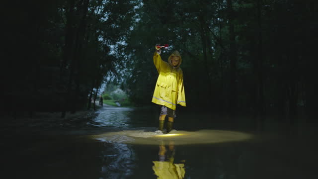 SLO MO Smiling Young Boy in Yellow Raincoat Walking in Flooded Road with Flashlight