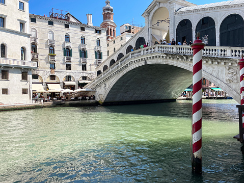 Stock photo showing close-up view of the Rialto Bridge (Ponte di Rialto) spanning the Grand Canal, with wooden boat docks and red and white striped canal poles (pali di casada), on a sunny Summer's day.