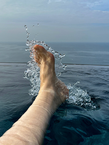 Stock photo showing close-up view of bare foot kicking and splashing water on surface of infinity swimming pool.