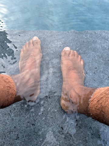 Stock photo showing close-up, elevated view of bare feet resting on top step of swimming pool visible through clear water.