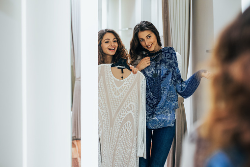 Two friends trying on clothes in a dressing room. Female friends choosing dresses in luxury boutique. Girlfriends get dressed up in the fitting room and having great time together.