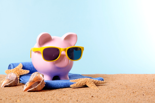 Pink piggy bank on a beach with sunglasses and beach towel.  Studio shot with plain blue background.  Space for copy.  Warm color and directional lighting are intentional.  Alternative version with white background shown below: