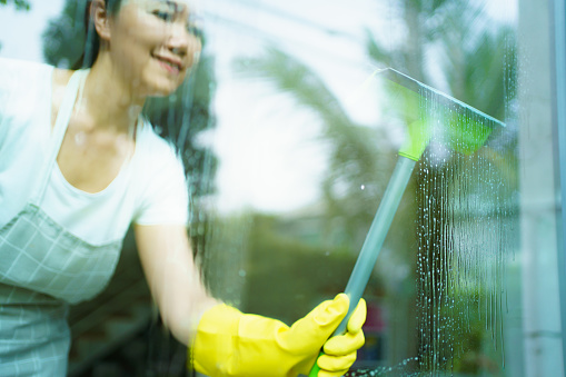 Cheerful happy Asian woman using a rubber squeegee cleaning a window close up, housewife wears a yellow rubber protective gloves while doing a chores or houseworks.