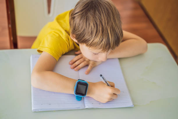 Little boy sitting at the table and looking smart watch. Smart watch for baby safety. The child makes school lessons, listening to music, calling friends stock photo