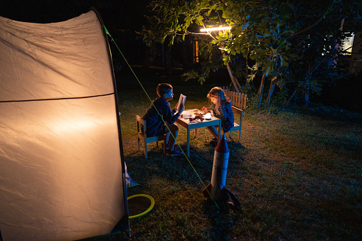 High angle view of children having fun on camping while sitting in front of tent, reading a book and drawing at night