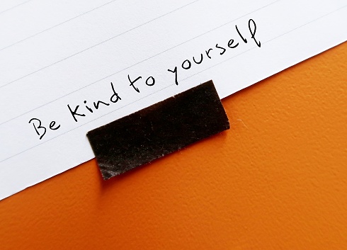 On orange background, handwritten text on note paper BE KIND TO YOURSELF, concept of self-kindness - be kind with understanding towards ourselves