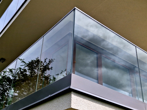 railing of a luxury house formed by glass panels fastened with metal stainless steel handles. gives an airy impression. polished metal cover on a greenhouse terrace window, reflection, block of flats