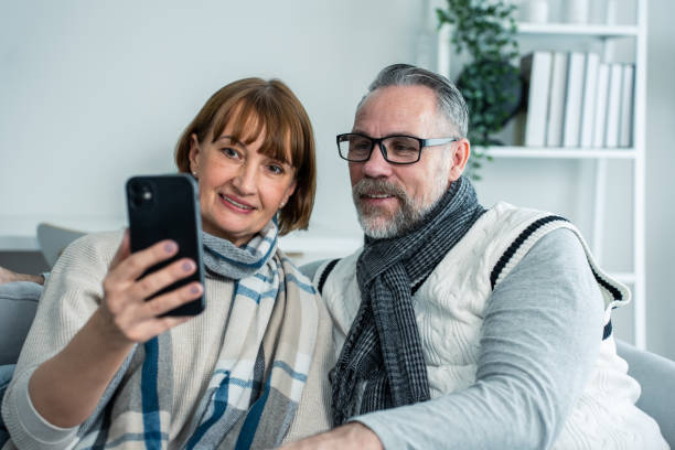 自宅のリビングルームで家族と白人の老夫婦のビデオ通話。携帯電話を使ってソファに座っている魅力的な年配の成熟した男性と女性が、家の親戚と仮想オンラインで通信します。 - senior women electronics people remote control ストックフォトと画像