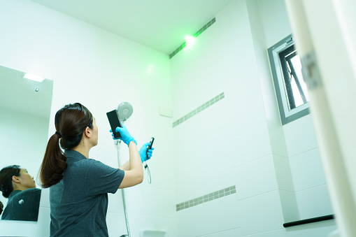 Professional Asian female insurance claim agent using a laser pointer pointing at the damaged ceiling and taking a photo on digital tablet and smartphone, female claim inspector inspecting.