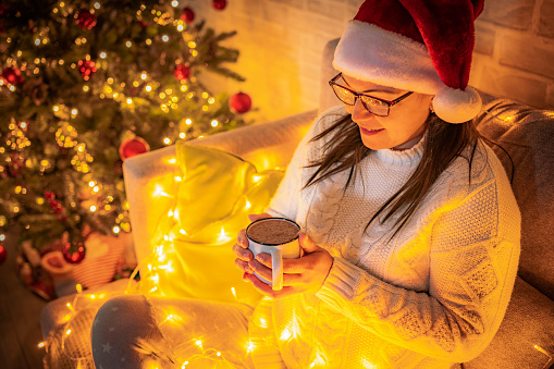 Woman wearing Santa hat sitting on sofa drinking hot chocolate. Defocused Christmas tree and string lights complete the composition. High resolution 42Mp indoors digital capture taken with Sony A7rII and and Zeiss Batis 40mm F2.0 CF lens