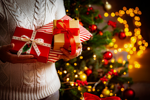 Close up view of woman's hands holding three Christmas presents with defocused Christmas tree and string lights at background.. High resolution 42Mp indoors digital capture taken with Sony A7rII and Sony FE 90mm f2.8 macro G OSS lens