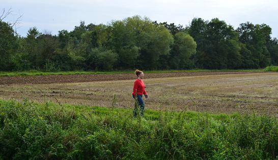 Leuven, Vlaams-Brabant, Belgium - August 31, 2021: tied up, red hair, eye wear, red shirt, blue jeans pants walking alone on a footpath along a farmers field on a cloudy day end summer