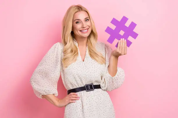 Photo of Photo portrait of attractive blonde woman holding hashtag number influencer dressed stylish white clothes isolated on pink color background