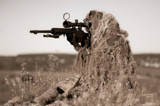 Special forces sniper in ghillie suit with a high-precision rifle with optic scope at combat position on the top of the rocky desert hillside