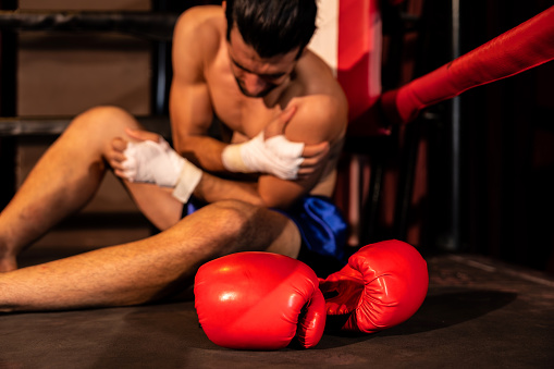 Caucasian boxer with pain and injury after intense boxing training or fighting match, sitting at the edge of ring. Physical injury in sport concept. Impetus