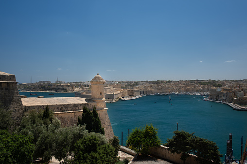 Valletta harbor in Malta photographed from above. You can see a tower, part of the city wall and trees in the foreground. In the background the sea with the harbor basin and part of the city.