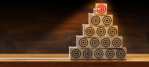 Ladder of success or goal concept. Symbol of an arrow going up a stack of wooden blocks with many brown targets and one red target being hit, above a wooden table or desk with copy space.