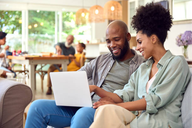 Casal em casa usando laptop para fazer compras ou reservar férias com a família de várias gerações em segundo plano - foto de acervo