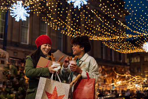 Two women enjoying a Christmas market on a cold December day in the city. They are eating churros while wrapped up warm and carrying gift bags.