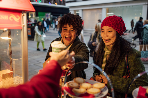 Waist-up shot of two women being passed a hot drink from a food and drink Christmas market on a cold December day in the city. They are wrapped up warm and talking to each other.
