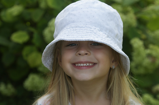 Lifestyle headshot portrait of beautiful, happy, white, Caucasian girl looking directly into the camera while smiling and wearing white summer floppy hat decorated with bandana in fresh flower colours. Shot on Canon EOS, photographed outdoors in nature with selective focus and shallow depth of field, sharp focus on girl's eyes, ISO 100.