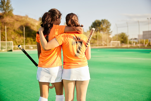 Two female teen field hockey players exit the field together as teammates in victory or defeat.