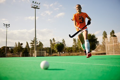 Stationary field hockey ball in the foreground, while a cheerful player approaches it in the background