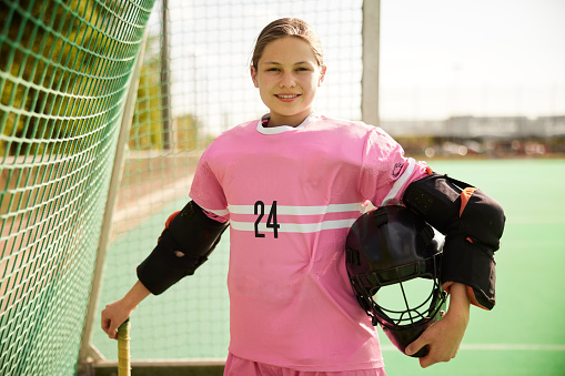 The teenage female field hockey goalkeeper exudes confidence as she poses for the camera, holding her field hockey stick in one hand and her helmet under the other arm, all while wearing a cheerful smile.