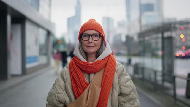 Portrait urban style happy middle aged older lady standing outside in city, trendy winter outfit bright orange hat and scarf