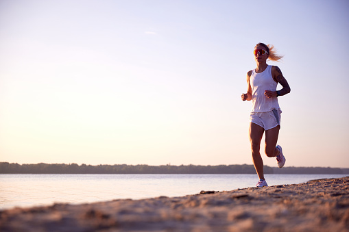 Full length portrait of young healthy woman in sunglasses, training, running at beach near river on sunrise. Morning fitness exercise in motion Concept of sport, recreation, healthy lifestyle, hobby