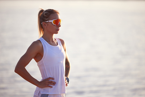 Portrait of active woman runner in sport clothes and sunglasses on seashore. Woman relaxing, resting after morning fitness routine. Concept of sport, recreation, healthy lifestyle, hobby.