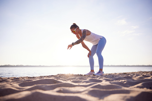 Full-lenght portrait of active woman runner in sport clothes checking athletic tracker on seashore. Woman resting after morning fitness routine. Concept of sport, recreation, healthy lifestyle
