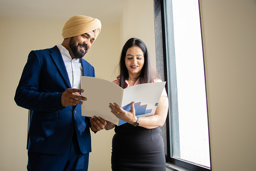 Indian sikh businessman wearing formal outfit discuss work with young female secretary holding file in corporate office.