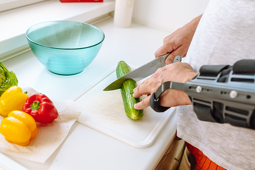 man with rigid elbow orthopedic holder with hinge, modern adjustable elbow orthosis, makes salad, from fresh vegetables, pepper cabbage, peels, cuts vegetables with kitchen knife in home kitchen