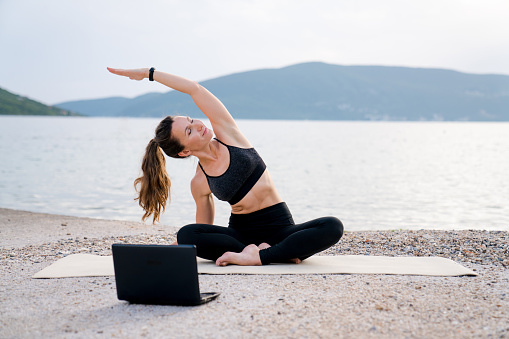 Yoga at sea beach online with laptop. Woman doing morning exercises for virtual class. Sport workout webinar on nature. Girl practicing fitness on wellness trip. Healthy lifestyle, activities outdoors