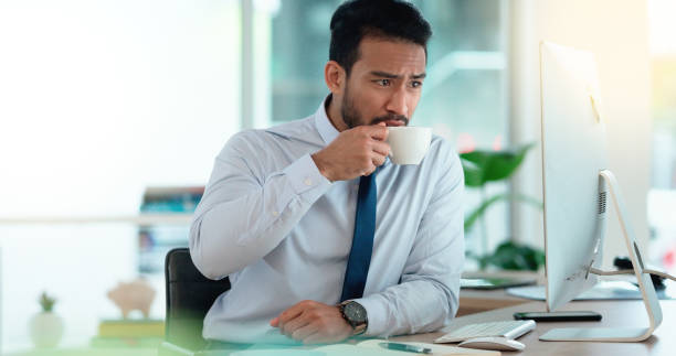 Exhausted businessman typing on a computer working and sipping coffee in a corporate office. A male employee sitting at a desk feeling sleepy and yawning tired from overworked job and drinking coffee