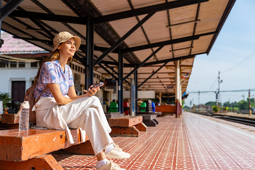 Caucasian woman tourist travel in Thailand on local train. Attractive girl using mobile phone with internet during tourist waiting for the train travel on railroad transportation on summer holiday vacations.