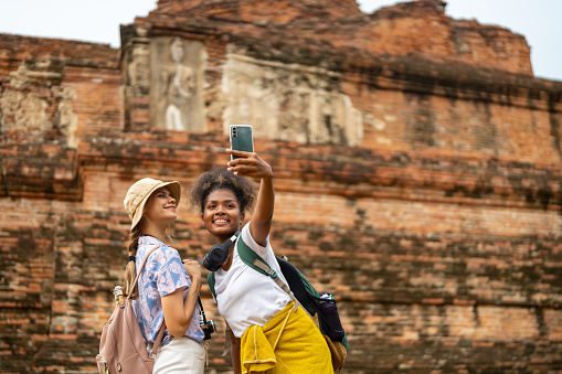Diversity woman friends using mobile phone taking selfie or vlogging together with architecture and ancient old ruin temple during travel famous place in Ayutthaya Province, Thailand on summer holiday vacation.