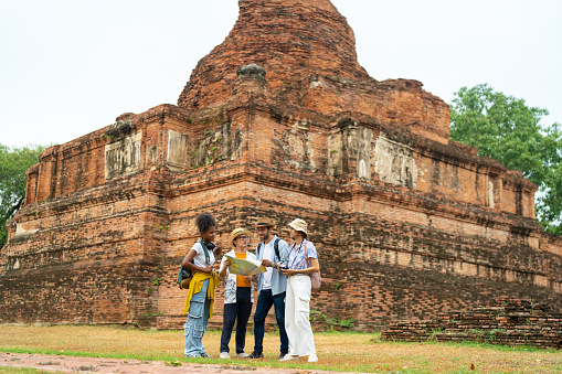 Group of Diversity people traveler travel Ayutthaya Province in Thailand on summer holiday vacations. Asian man local tour guide explaining about architecture and history of old ruin famous place temple to tourist.