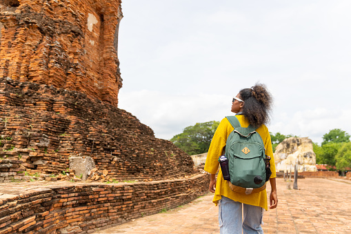 African woman tourist walking and looking at architecture around old ruin temple famous place during travel in Ayutthaya Province, Thailand on summer holiday vacation.