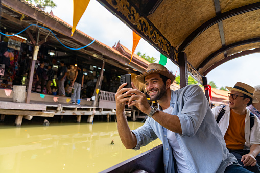 Group of Diversity people and local tour guide using mobile phone taking selfie or vlogging together during travel floating market by traditional wooden boat in canal in Ayutthaya Province, Thailand on summer holiday vacation.