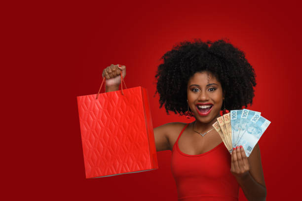 uma mulher segura sacolas de compras e dinheiro brasileiro, uma adolescente sorri e olha para a câmera, usando um vestido vermelho contra um fundo vermelho - real people young women women beautiful - fotografias e filmes do acervo