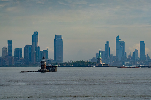 A small lighthouse and the statue of liberty with the Manhattan skyline in the background, on a hazy day.
