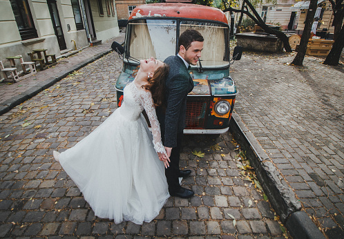 Wedding couple laughs in the old city. Colorful hippie bus in ancient autumn town on background. Rustic bride with hair down and groom in grey suit and bow tie. Romantic love in vintage atmosphere.
