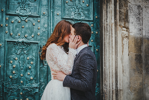 Wedding couple kisses near the vintage green door. Stone walls in ancient town background. Rustic bride with hair down in lace dress and groom in grey suit and bow tie. Tender embrace. Romantic hug.