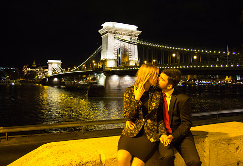 Couple in love is kissing and hugging with a view on Budapest Chain bridge. Night city lights in european city. Man and woman silhouettes near river panoramic view. Stylish fashion photo.
