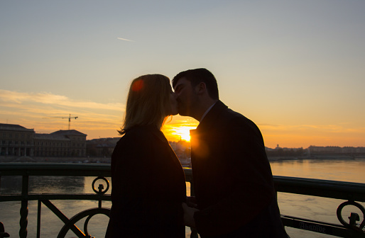 Couple in love is standing on the liberty bridge over the river Danube in Budapest. Sunrise in the big city. Dark silhouettes of man and woman holding hands in urban landscape. Sun kiss.