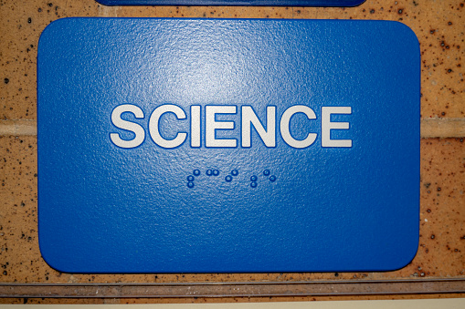 Blue science sign with braille, on brick wall at door to school office.
