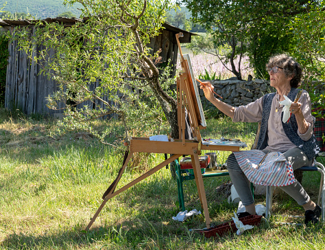 A happy senior woman wearing a wide-brimmed hat is sitting outdoors in a park, in front of a canvas on an easel.  She is holding a palette in one hand and a paint brush in the other.  The background is lush, green foliage.